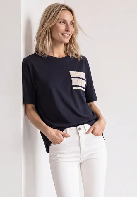 Eine lächelnde Frau lehnt in weißer Jeans und schwarzem Shirt mit Brusttasche an einer Wand.