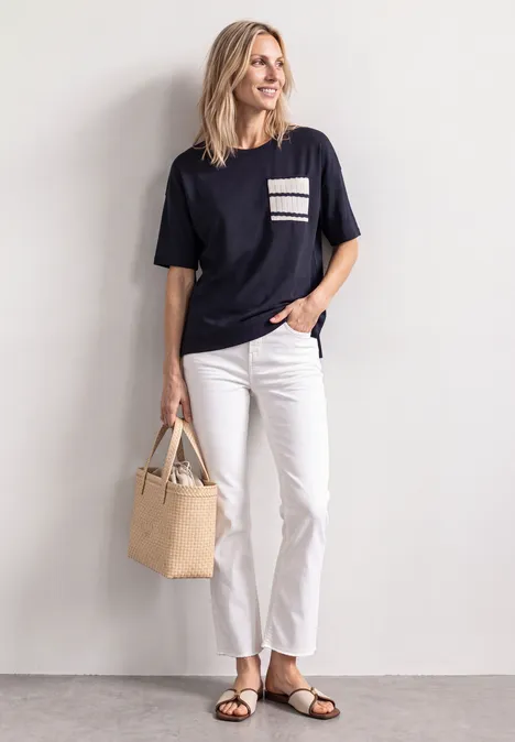 Frau in legerem Outfit: dunkles T-Shirt mit gestreifter Brusttasche, weiße Hose, Sandalen und Strohtasche.