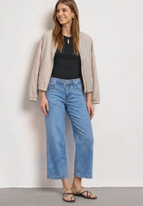 Dropped Shoulder Shirt mit Cut-Out Black Eine Frau steht vor weißem Hintergrund in blauen, weiten Cropped-Jeans, schwarzem Top, beigem Blazer und Sandalen.