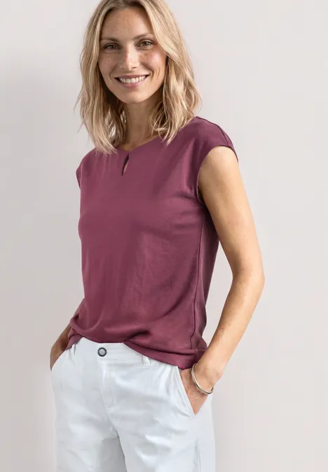 Eine Frau trägt ein ärmelloses violettes Top und weiße Hosen. Sie steht mit Händen in den Taschen.