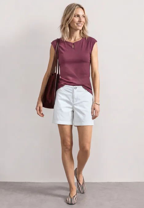Frau trägt bordeauxfarbenes Top, weiße Shorts und Flip-Flops, hält große Tasche vor neutralem Hintergrund.