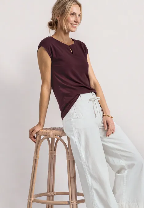 Femme appuyée sur un tabouret en bois, porte un haut bordeaux à manches courtes et un pantalon large clair.