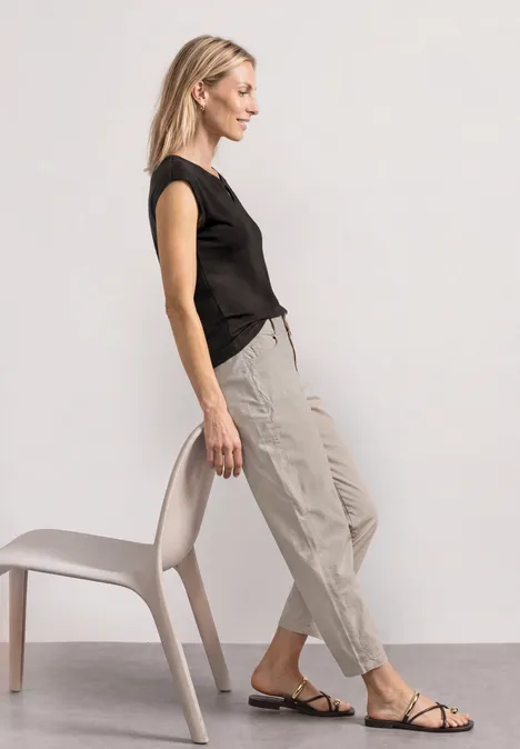 Dropped Shoulder Shirt mit Cut-Out black coffee Frau in schwarzem Shirt und beigen Stoffhosen lehnt sich seitlich an einen Stuhl, trägt flache Sandalen.
