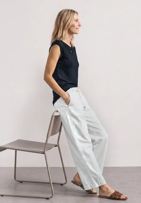 Dropped Shoulder Shirt mit Cut-Out darkest blue Eine Frau in schwarzem Top, weiten weißen Hosen und Sandalen steht seitlich neben einem minimalistischen Stuhl.