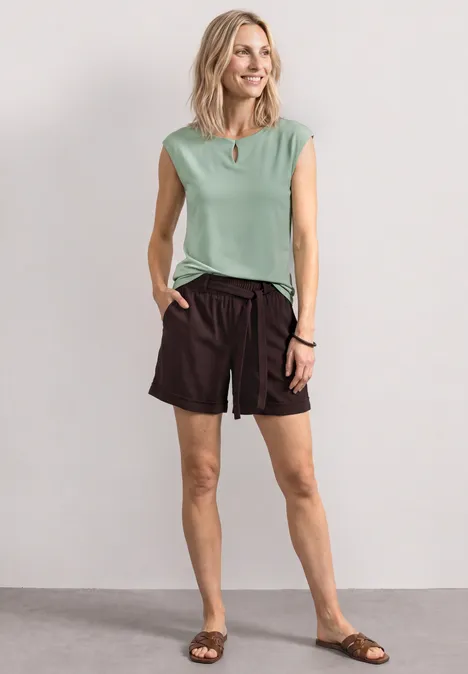 Frau im mintgrünen Top, schwarzen Shorts und Sandalen steht locker vor neutraler Wand.