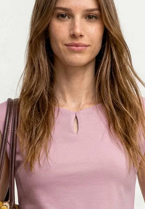 Frau in rosa Oberteil mit Schlüsselloch-Ausschnitt, brauner Schultertasche, lange braune Haare, weißer Hintergrund
