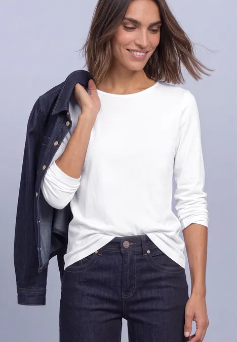 Basic Langarm Shirt in Unifarbe White Frau in weißem Langarmshirt und dunkler Jeansjacke über der Schulter, trägt dunkle Jeans und lächelt.