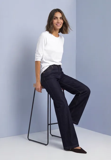 Basic Langarm Shirt in Unifarbe White Frau sitzt seitlich auf Hocker, trägt weiße Bluse, weite dunkle Hose und schwarze Schuhe.