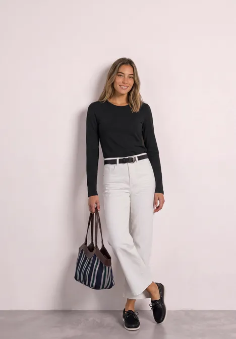 Jednokolorowa koszulka Basic z długim rękawem Black Kobieta w czarnej bluzce z długim rękawem, białych szerokich spodniach, czarnym pasku, pasiastą torbą i czarnych mokasynach, uśmiechnięta.