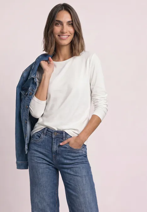 Jednokolorowa koszulka Basic z długim rękawem off white Uśmiechnięta kobieta w białej bluzce i dżinsach z kurtką przewieszoną przez ramię