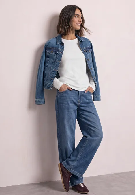 Jednokolorowa koszulka Basic z długim rękawem off white Kobieta w casual denimowym zestawie: biała bluzka, jeansy i jeansowa kurtka, uśmiechająca się oparta o jasną ścianę.