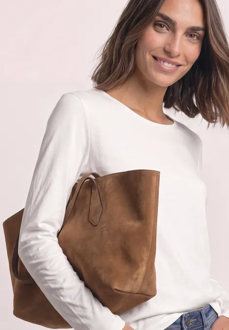 Jednokolorowa koszulka Basic z długim rękawem off white Kobieta w białej bluzce z długim rękawem i dżinsach trzyma pod pachą brązową zamszową torbę.
