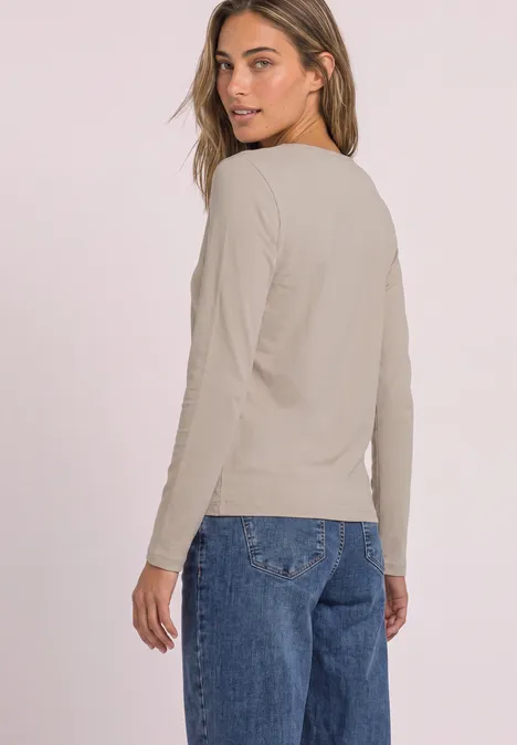 T-shirt basique à manches longues, couleur unie clay sand Frau trägt beiges Langarmshirt und blaue Jeans, schaut über die Schulter vor hellem Hintergrund.
