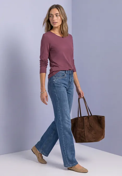 Femme en haut violet, jean bleu et chaussures beiges, qui porte un grand sac en daim marron.
