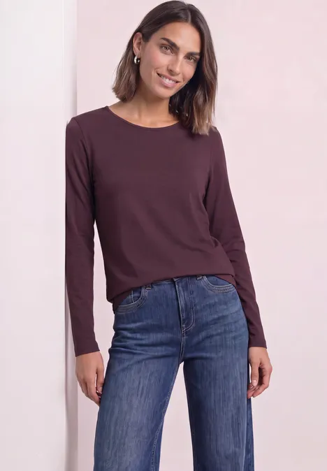 Jednokolorowa koszulka Basic z długim rękawem rustic rouge Kobieta w bordowej bluzce z długim rękawem i niebieskich dżinsach, uśmiechnięta, z półdługimi włosami i kolczykami.