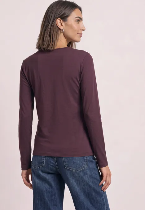 Jednokolorowa koszulka Basic z długim rękawem rustic rouge Kobieta plecami do aparatu, ubrana w bordową bluzkę z długim rękawem i dżinsy na jasnym tle.
