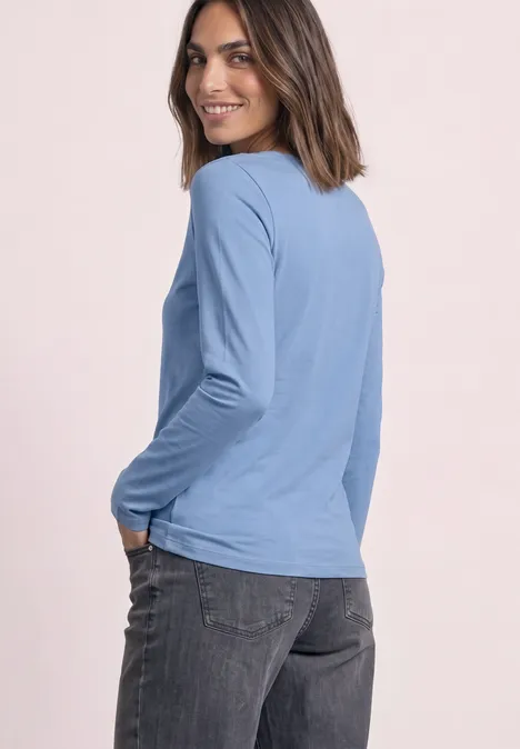Jednokolorowa koszulka Basic z długim rękawem northern blue Kobieta w jasnoniebieskiej bluzce z długim rękawem i ciemnych dżinsach, uśmiechnięta na różowym tle.