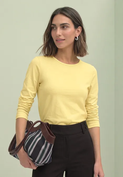Vrouw in gele trui met lange mouwen en donkere broek, met gestreepte handtas.