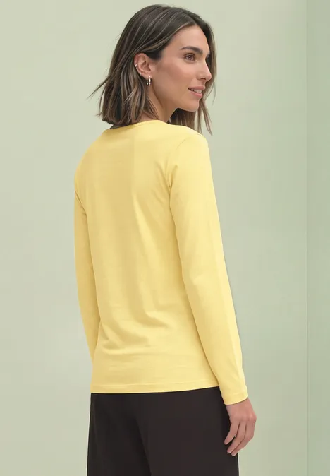 Vrouw in geel langarmshirt zijwaarts staand tegen een lichtgroene achtergrond.
