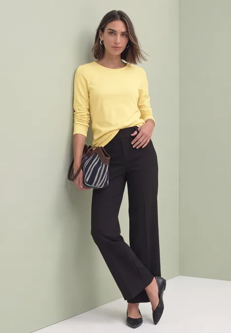 Vrouw in geel shirt met lange mouwen, zwarte broek en zwarte schoenen, met schoudertas voor een lichte achtergrond.