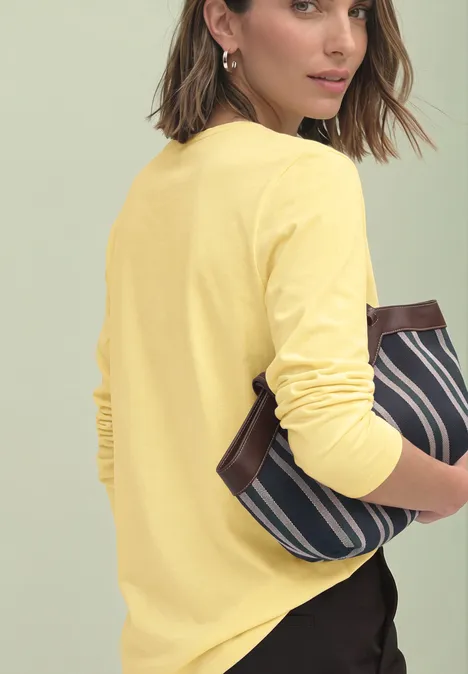 Een vrouw in gele longsleeve houdt een blauw-wit gestreepte clutch vast en kijkt over haar schouder.