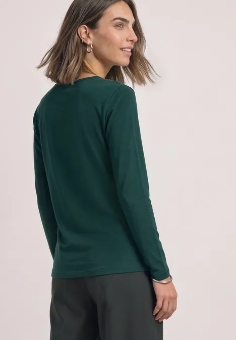 Basic Langarm Shirt in Unifarbe shadow teal Eine Frau im dunkelgrünen Langarmshirt und schwarzer Hose steht seitlich und blickt über die Schulter.