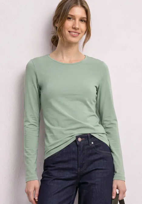 Jednokolorowa koszulka Basic z długim rękawem eucalyptus green Kobieta w miętowej bluzce z długim rękawem i ciemnych jeansach, stojąca i uśmiechnięta na jasnym tle.