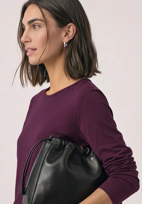 Basic longsleeve berry maroon Vrouw in paars shirt met kort haar opzij, houdt een zwarte leren buideltas vast.
