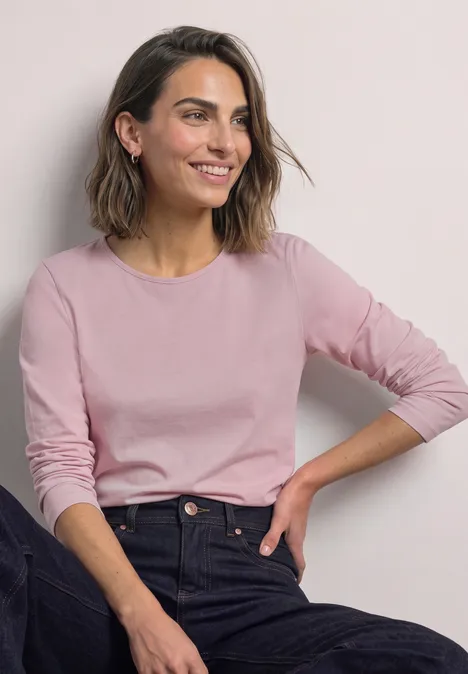 Eine lächelnde Frau in rosa Langarmshirt und dunkler Jeans sitzt entspannt vor neutralem Hintergrund.