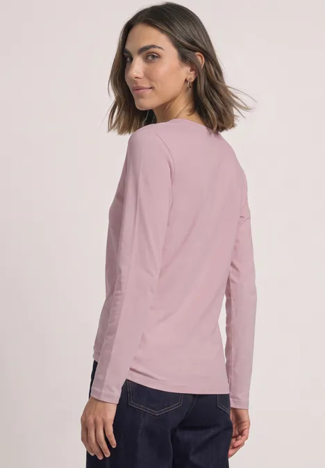 Eine Frau in rosa Langarmshirt und dunkler Hose steht seitlich und schaut über die Schulter.