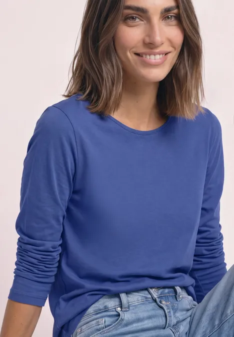 Junge Frau sitzt lächelnd vor hellem Hintergrund, trägt blaues Langarmshirt und Jeans.