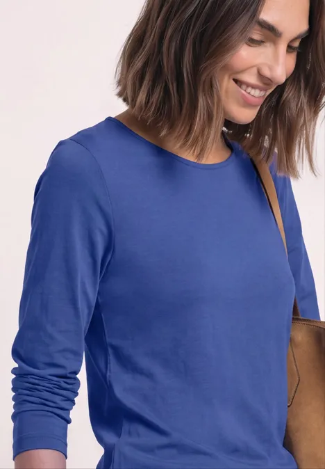 Eine lächelnde Frau in blauem Langarmshirt mit Umhängetasche vor hellem Hintergrund.