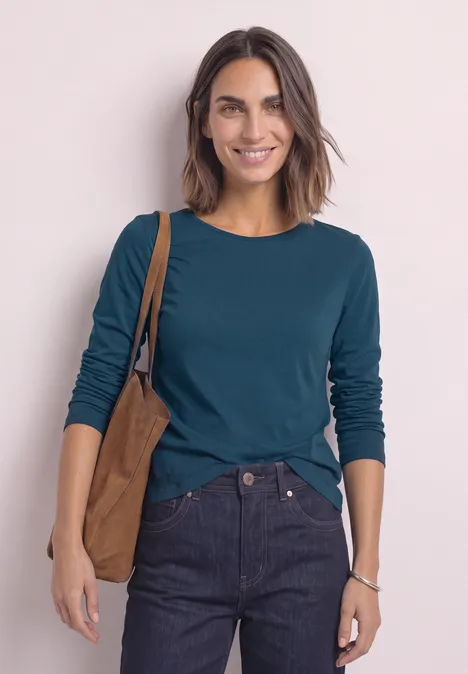 T-shirt basique à manches longues, couleur unie blue petrol Frau mit dunklen Haaren in dunkelgrünem Langarmshirt und Jeans, lächelnd mit brauner Tasche