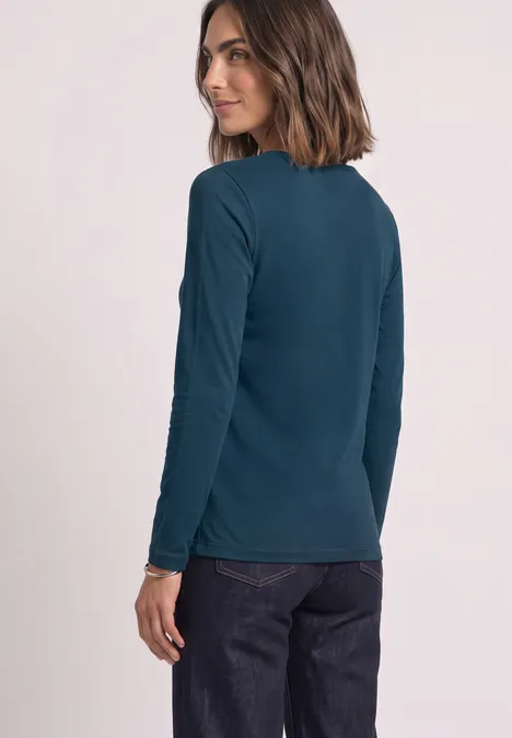 T-shirt basique à manches longues, couleur unie blue petrol Frau mit schulterlangen braunen Haaren trägt ein petrolfarbenes Langarmshirt und dunkle Hose, schaut über die Schulter.