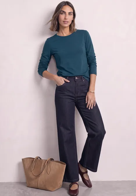 T-shirt basique à manches longues, couleur unie blue petrol Frau in tealfarbenem Shirt, schwarzen Jeans und braunen Loafers, daneben eine große Tasche vor hellem Hintergrund.