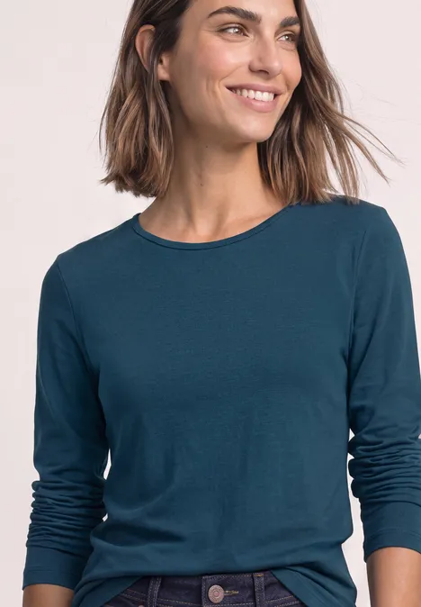 T-shirt basique à manches longues, couleur unie blue petrol Eine lächelnde Frau trägt ein petrolfarbenes Langarmshirt und Jeans vor hellem Hintergrund.