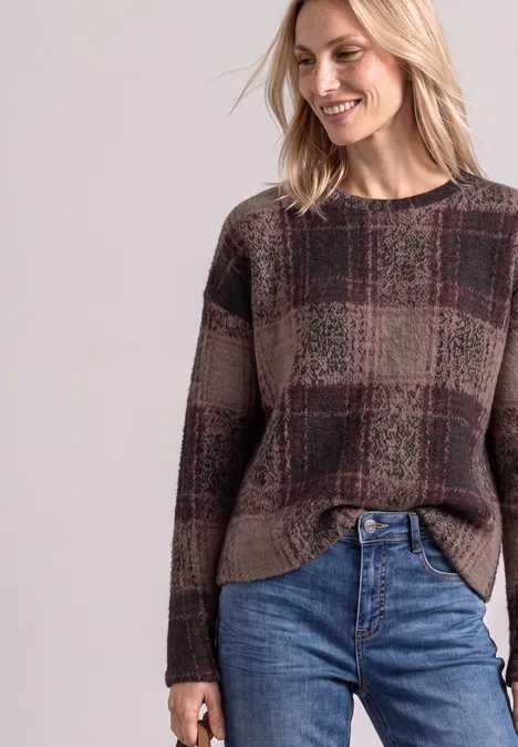 Femme souriante portant un pull à carreaux tons terre et un jean, sur fond neutre.
