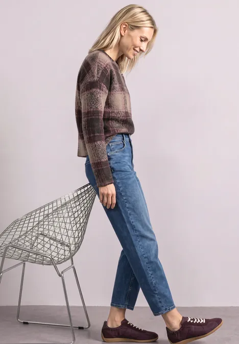 Femme en pull à carreaux et jean, appuyée de manière décontractée sur une chaise en métal.