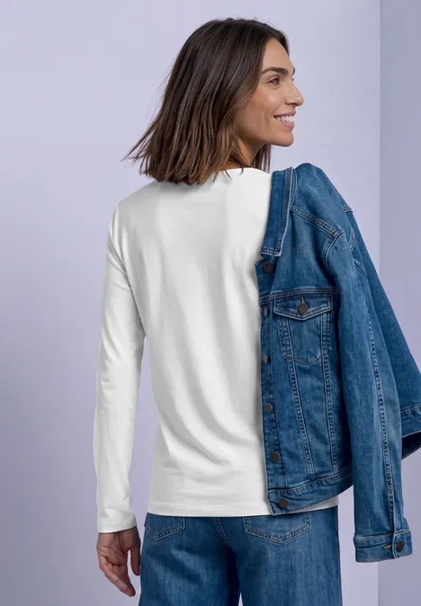 Koszulka z długim rękawem i dekoltem w kształcie kwadratu off white Kobieta w białej bluzce z długim rękawem trzyma jeansową kurtkę na ramieniu i uśmiecha się na tle jasnofioletowym.