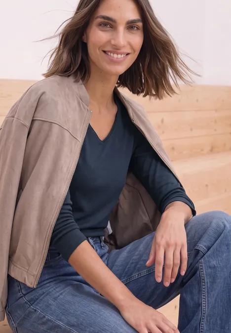Shirt met lange mouwen en vierkante halslijn darkest blue Frau mit offener Wildlederjacke über den Schultern, dunkelblauem Pullover und Jeans, lächelt vor Holzpaneel.
