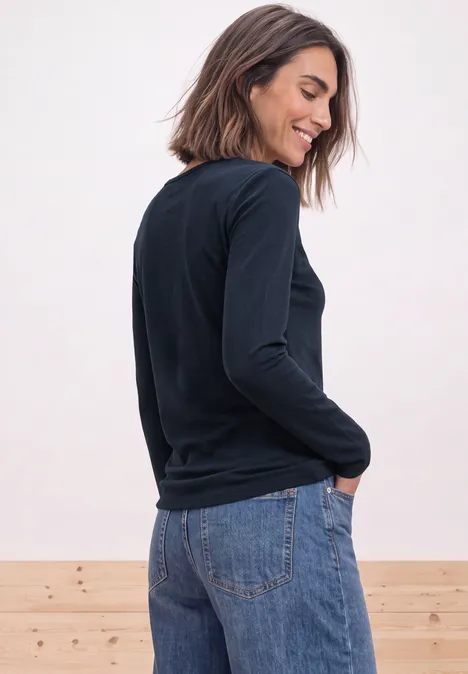 Shirt met lange mouwen en vierkante halslijn darkest blue Eine lächelnde Frau im schwarzen Pullover und Jeans steht seitlich vor einer Holzwand und schaut zurück.