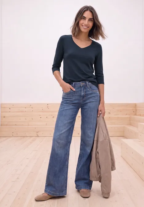 Shirt met lange mouwen en vierkante halslijn darkest blue Frau in navyblauem V-Shirt und weiten Jeans, hält beige Jacke vor hellem Holz-Wand und -Boden.