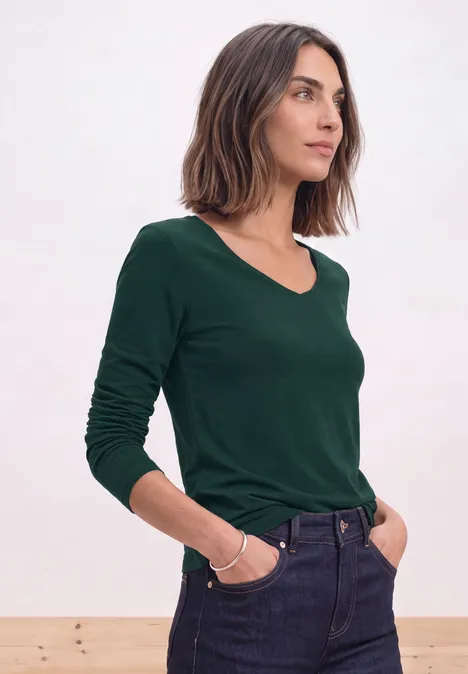 Femme aux cheveux mi-longs en haut vert à manches longues et jean, debout de profil, mains dans les poches.