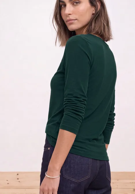 Femme tournée vers la caméra, porte un haut vert à manches longues et un jean foncé