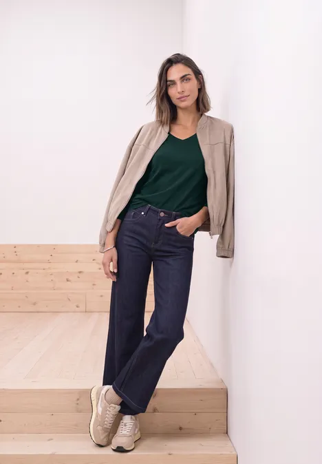 Femme en jean, top vert et baskets claires appuyée sur un mur blanc, debout sur des marches en bois clair.