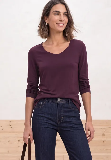 Shirt met lange mouwen en vierkante halslijn berry maroon Frau in lila V-Ausschnittshirt und dunklen Jeans, lächelnd mit Tasche vor hellem Hintergrund.