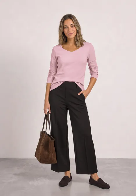 Frau in rosa V-Pullover, schwarzen weiten Hosen, brauner Tasche, schwarzen Slippern vor hellem Hintergrund.