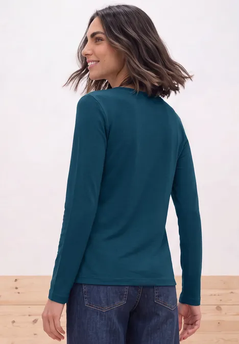 Frau von hinten in petrolfarbenem Langarmshirt und Jeans, schulterlange dunkle Haare, seitlich lächelnd.