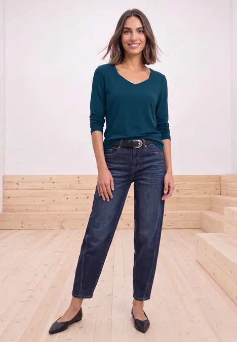 Frau mit petrolfarbenem Langarmshirt, dunklen Jeans, Gürtel und schwarzen Ballerinas auf hellen Holzstufen.