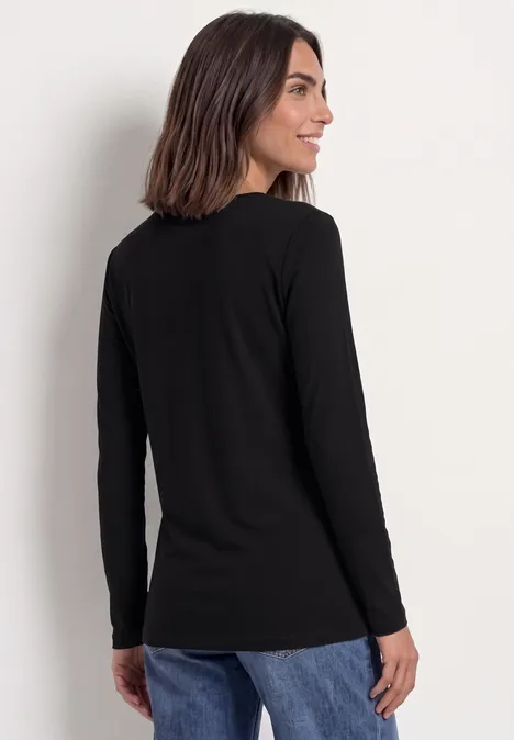Femme aux cheveux mi-longs foncés, vêtue d'un tee-shirt noir à manches longues et d'un jean bleu, de profil devant un mur blanc.